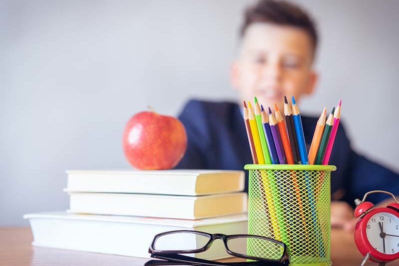Apple sitting on books alongside pencils, reading glasses, and a small clock